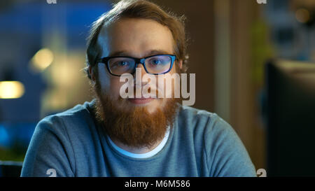 Young Bearded Creative Man With Glasses Thinks on a Problem While Working on His Personal Computer. He Looks Smilingly Into the Camera. Stock Photo