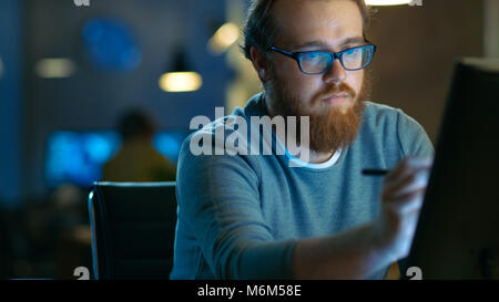Talented Young Concept Artist Drawing on a Digital Pen Display Tablet While Sitting at His Working Place in Modern Loft Office. Late at Night. Stock Photo