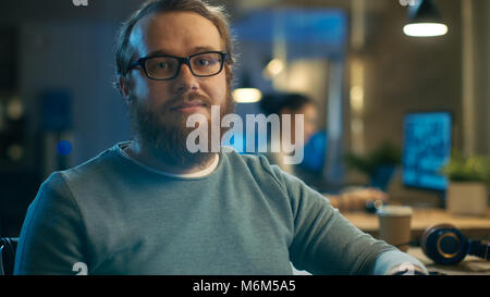 Young Bearded Creative Man With Glasses Thinks on a Problem While Working on His Personal Computer. He Looks Smilingly Into the Camera. Stock Photo