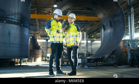 Male and Female Industrial Engineers in Hard Hats Discuss New Project while Using Tablet Computer. They Make Showing Gestures. Heavy industry. Stock Photo