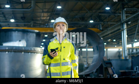 Female Industrial Worker in the Hard Hat Uses Mobile Phone While Walking Through Heavy Industry Manufacturing Factory. In the Background Various Metal Stock Photo