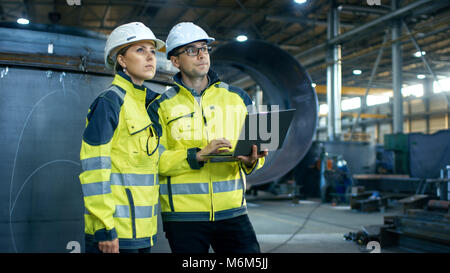 Male and Female Industrial Engineers in Hard Hats Discuss New Project while Using Laptop. They Make Showing Gestures.They Work at the Heavy Industry Stock Photo