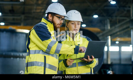 Male and Female Industrial Engineers in Hard Hats Discuss New Project while Using Laptop. They Make Showing Gestures.They Work at the Heavy Industry Stock Photo