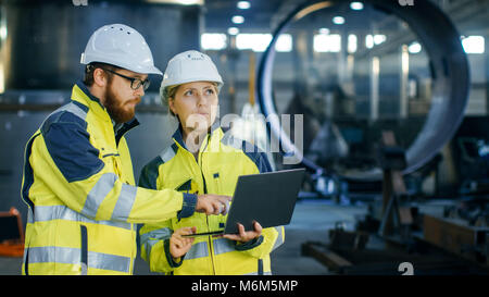 Male and Female Industrial Engineers in Hard Hats Discuss New Project while Using Laptop. They Make Showing Gestures.They Work in a Heavy Industry Stock Photo
