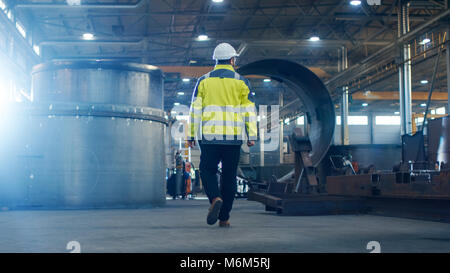Industrial Engineer in Hard Hat Wearing Safety Jacket Walks Through Heavy Industry Manufacturing Factory with Various Metalworking Processes. Stock Photo