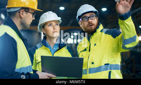 Male and Female Industrial Engineers Talk with Factory Worker while Using Laptop. They Work at the Heavy Industry Manufacturing Facility. Stock Photo