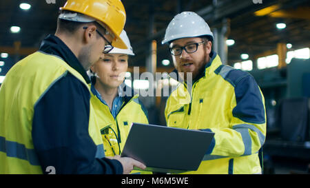 Three Industrial Engineers Talk with Factory Worker while Using Laptop. They Work at the Heavy Industry Manufacturing Facility. Stock Photo