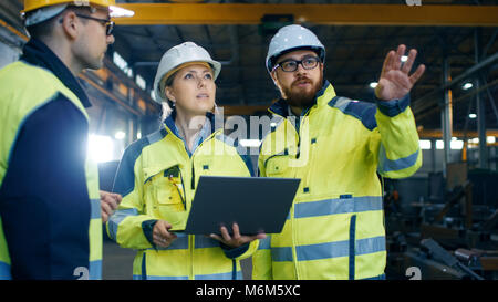 Male and Female Industrial Engineers Talk with Factory Worker while Using Laptop. They Work at the Heavy Industry Manufacturing Facility. Stock Photo