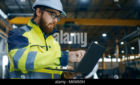 Industrial Engineer in Hard Hat Wearing Safety Jacket Uses Touchscreen Laptop. He Works at the Heavy Industry Manufacturing Factory. Stock Photo