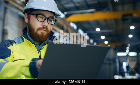 Industrial Engineer in Hard Hat Wearing Safety Jacket Uses Touchscreen Laptop. He Works at the Heavy Industry Manufacturing Factory. Stock Photo