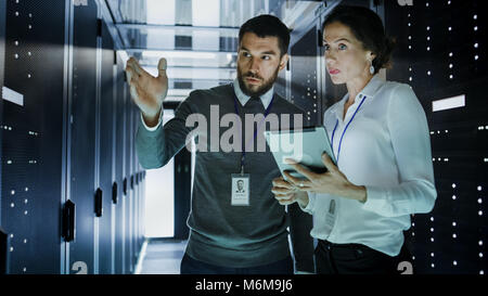 Two IT Engineers in Data Center / Server Room, Talking, one of them Pointing Forward, Other Holds Tablet Computer. Stock Photo