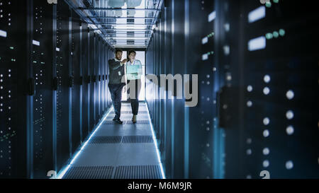 Female IT Technician and Male Server Engineer Talk and Discuss. They are in Working Data Center full of Rack Servers. Woman Holds Laptop. Stock Photo