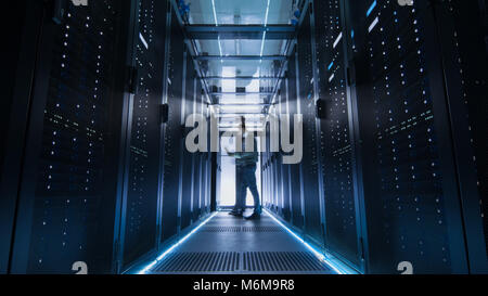 Male IT Engineer Walking and Working in Data Center Corridor. She is Holding Tablet Computer. He is Blurred in Motion. Stock Photo