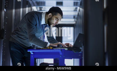 IT Engineer with Tool Cart Working on a Laptop Computer, he Deploys new software. He Stands at a Corridor of a Large Data Center Full of Rack Servers. Stock Photo