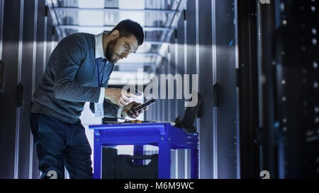 IT Engineer with Tool Cart Working on a Laptop Computer, he Deploys new software. He Stands at a Corridor of a Large Data Center Full of Rack Servers. Stock Photo