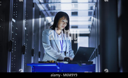 Asian Female IT Engineer Working on a Laptop on Tool Cart, She Scans Hard Drives.  She's in a Big Data Center Full of Rack Servers. Stock Photo