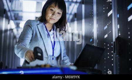 Asian Female IT Engineer Working on a Laptop on Tool Cart, She Scans Hard Drives.  She's in a Big Data Center Full of Rack Servers. Stock Photo