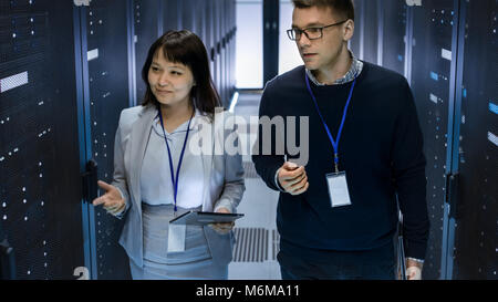 Caucasian Male and Asian Female IT Technicians Walking through Corridor of Data Center with Rows of Rack Servers. They Have Discussion Stock Photo