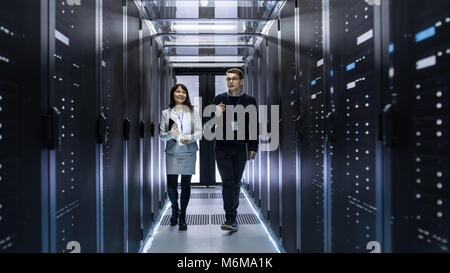 Caucasian Male and Asian Female IT Technicians Walking through Corridor of Data Center with Rows of Rack Servers. They Have Discussion Stock Photo