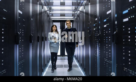 Caucasian Male and Asian Female IT Technicians Walking through Corridor of Data Center with Rows of Rack Servers. They Have Discussion Stock Photo