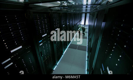 Security Camera view of a Hooded Hacker in Data Center, With His Laptop He Connects to One of The Rack Servers, Commits Crime. Stock Photo