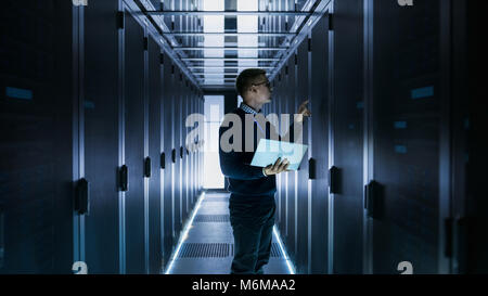 Male IT Engineer Works on a Laptop in front of Server Cabinet at a Big Data Center. Rows of Rack Servers are Seen. Stock Photo