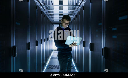 Male IT Engineer Works on a Laptop in front of Server Cabinet at a Big Data Center. Rows of Rack Servers are Seen. Stock Photo