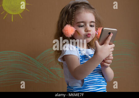 Close-up of a beautiful little girl smiling with a funny face makes selfie on a background of painted sun and white clouds Stock Photo