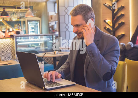 Indoor shot of young man sitting at a cafe table using laptop and talking on mobile phone. Handsome caucasian male sitting at restaurant and working. Stock Photo