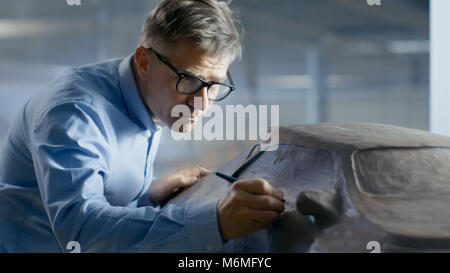 Professional Automotive Designer with Rake Sculpts Futuristic  Car Model from Plasticine Clay. He Works in a Special Studio Located In a Car Factory Stock Photo