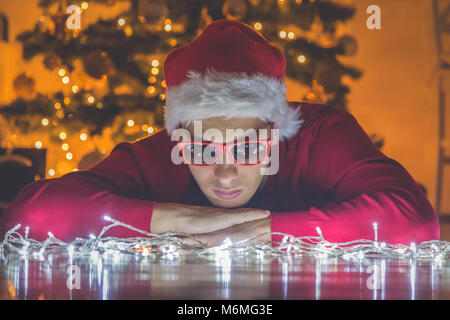 Thoughtful man with Santa hat lying on the floor and looking at Christmas lights. Christmas tree in the background Stock Photo