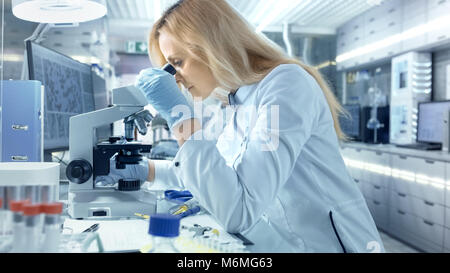 Female Research Scientist Looks at Biological Samples Under Microscope. She and Her Colleagues Work in a Big Modern Laboratory. Stock Photo