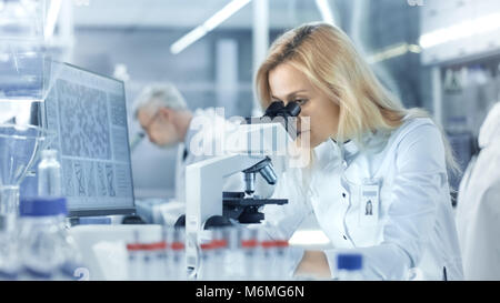 Female Research Scientist Looks at Biological Samples Under Microscope. She and Her Colleagues Work in a Big Modern Laboratory/ Medical Centre. Stock Photo