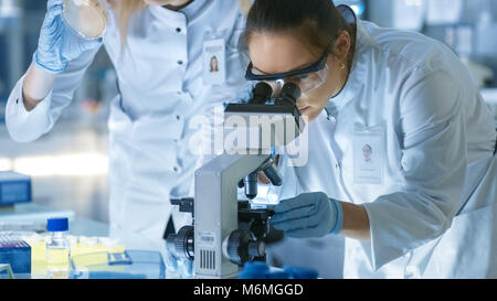 Medical Research Scientist Drops Sample on Slide and Her Colleague Examines it Under Microscope. They Work in a Modern Laboratory. Stock Photo