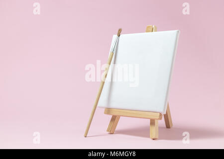 Blank art canvas on easel and paintbrush against pastel pink rose background. Artist minimal concept. Space for copy. Stock Photo