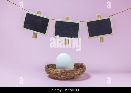 White egg in nest and clothespins with small blackboards hanging above against pastel pink background minimal easter concept. Stock Photo