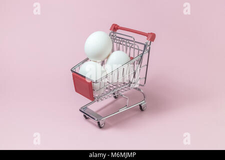 Shopping trolley model full of white eggs against pastel pink rose background minimal concept. Space for copy. Stock Photo