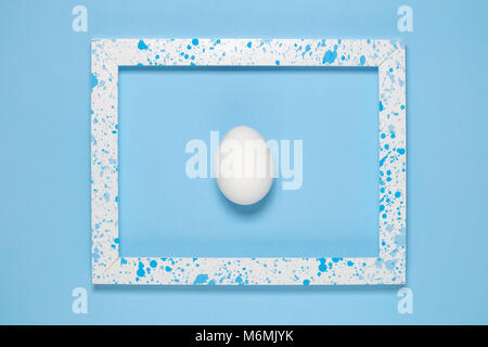 Flat lay of white egg in wooden frame colored with blue paint splatters on blue background minimal concept. Stock Photo
