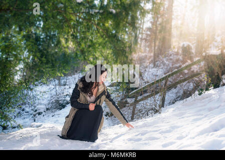 Young brunette woman, dressed in a winter jacket and  black skirt, in a squat position, reaching for snow, on a sunny day, in Blaubeuren, Germany. Stock Photo