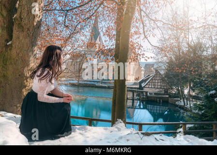 Young attractive woman, in a squat position, enjoying the view of the Blautopf spring and antique German architecture, in Blaubeuren, Germany. Stock Photo