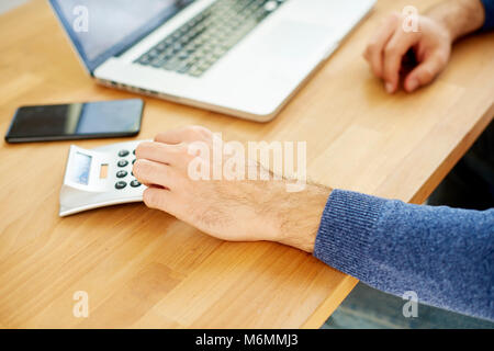 Close-up of businessman's hand counting on calculator while sitting at desk in front of laptop. Focus on foreground. Stock Photo