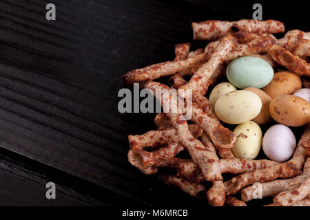 Easter nest made from edible crisps that look like sticks, and candy covered chocolate eggs. Birds nest on a black wooden table with copy space Stock Photo