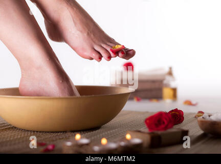 Female feet with drops of water, spa bowl, towels, flowers, candles, sea salt and bottle with essential oil on white background. Foot spa concept. Stock Photo