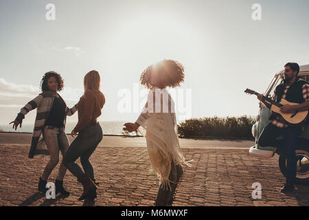 Group of friends on roadtrip dancing and having fun. Man playing guitar and women dancing along the road. Stock Photo