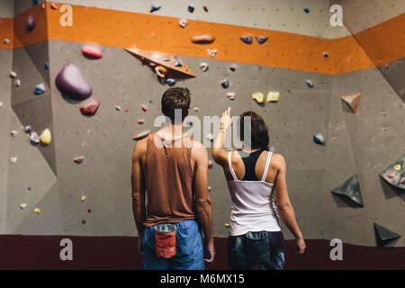 Rear view of female instructor giving instructions to a man on wall climbing. Man learning the art of rock climbing at an indoor climbing centre. Stock Photo