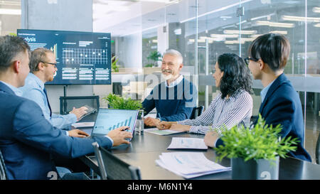 Senior Executive Explains Company's Vision and Potential to His Employees. They are Sitting at Big Table in Meeting Room. TV Screen on the Wall Stock Photo