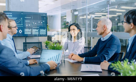 Diverse Team of Executives Have Meeting in a Conference Room. They Share Opinions, Statistics Show on Laptops and Tablets. In the Background TV Screen Stock Photo