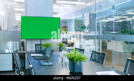 Modern Empty Meeting Room with Big Conference Table with Various Documents and Laptops on it, on the Wall Big TV with Green Chroma Key Screen. Stock Photo