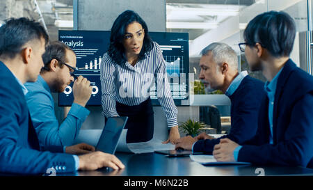 Chief Female Executive Leans and Spreads Project Blueprints on the Table Showing them to Her Colleagues. In the Background Pie Charts and Graphs Stock Photo