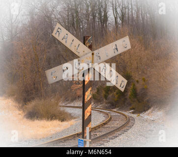 A vintage rail road crossing sign is viewed as the tracks curve around a bend to the left. Image was taken on a bleak winter day with leafless trees . Stock Photo